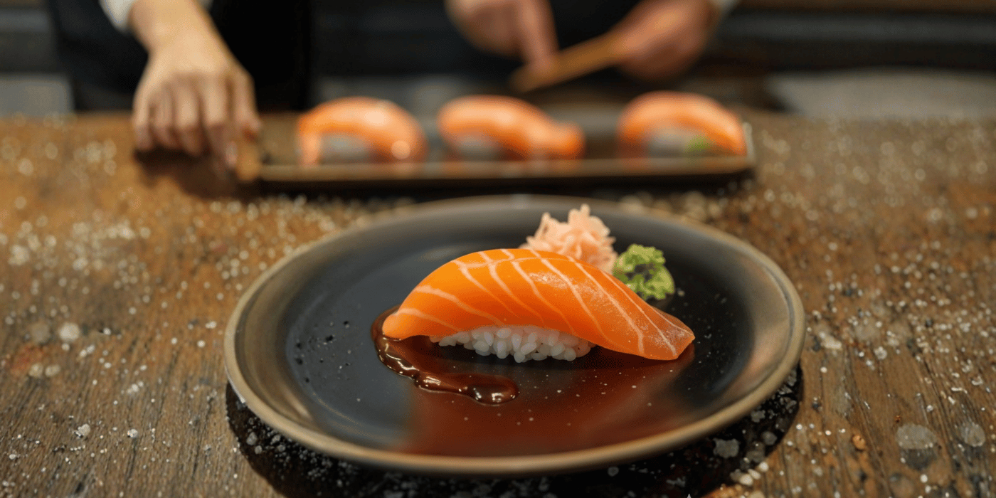 Plano cenital limpio de tres nigiri clásicos (salmón, atún, lubina) en tabla de madera, con palillos a un lado y fondo neutro. Capta “protagonismo del nigiri” desde el primer segundo.