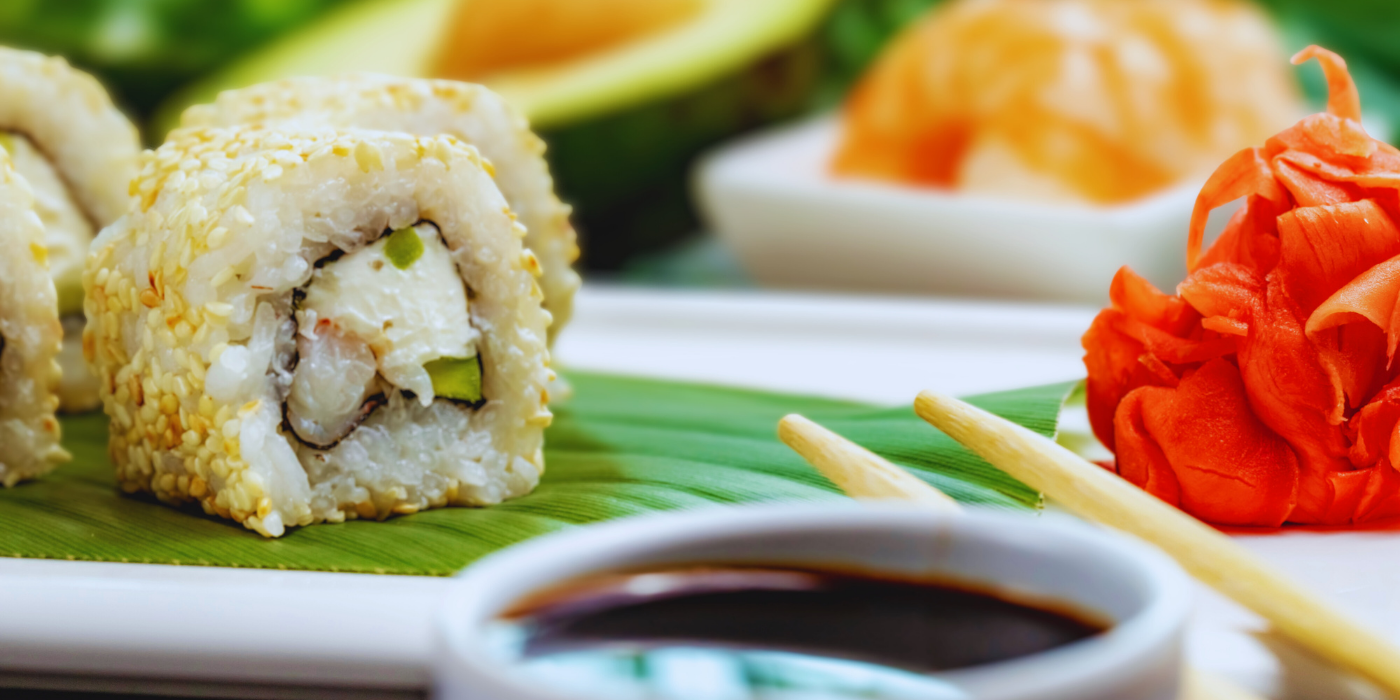 Primer plano horizontal de uramaki de salmón rebozado en sésamo, con gari y shōyu en primer término sobre hoja y cerámica clara. Paleta fresca y luminosa que introduce la ruta fría de primavera del Menú Infinity antes de pasar a nigiri y crujientes.
